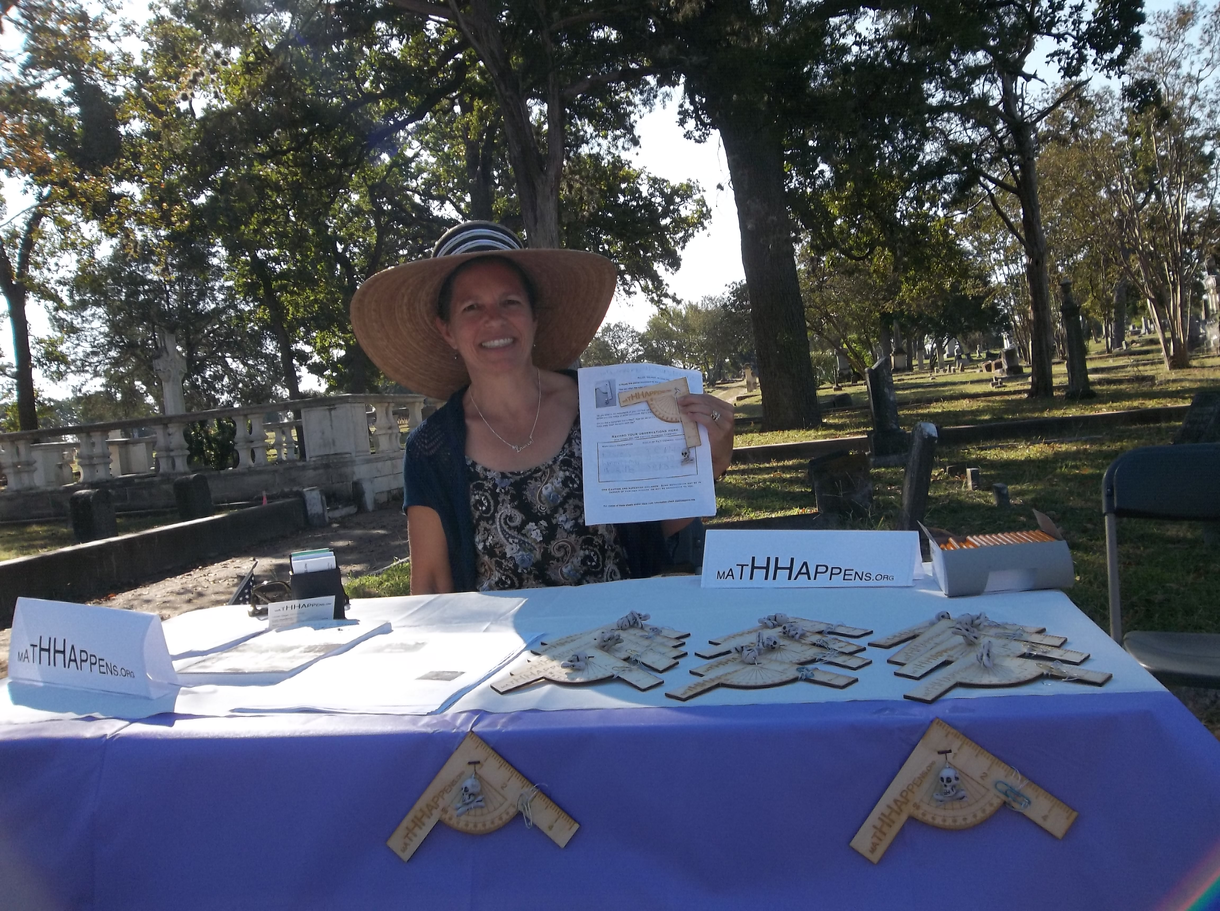 A woman in a sunhat sits at a table filled with wooden measurement devices. She is holding up an information sheet and smiling broadly
