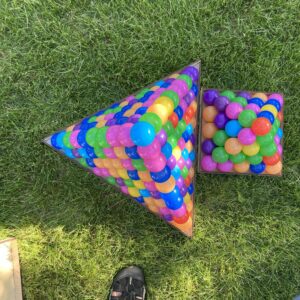 square (base 5) and triangular (base 10) pyramids made from multi-colored spheres inside wooden frames. They rest on a shaded patch of grassy ground