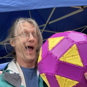 A man holds a large 3D shape made of purple pentagons and rectangles, and yellow triangles, while making a silly face with long, windblown hair in a selfie.