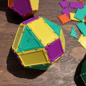 A 3D shape constructed from yellow rectangles, purple squares, and equilateral green triangles sits on a wooden table