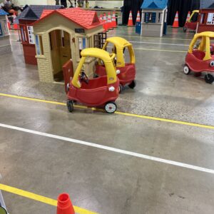 Several red and yellow plastic ride-in cars for toddlers are parked in the middle of a large indoor concrete floor, with toddler sized playhouses scattered about, yellow and white lines indicating roadways, and a large number of full-sized traffic cones.