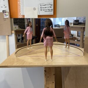 A Barbie stands on a platform, looking at herself in two mirrors at a 120° angle, seeing two reflections of herself.