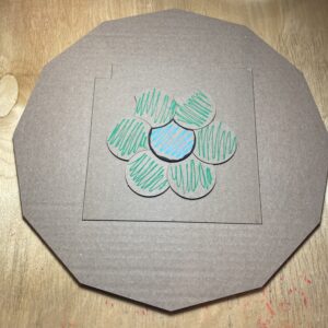 A blue teardrop-shaped tile is surrounded by six green ones on a cardboard surface. All of the tiles are congruent.